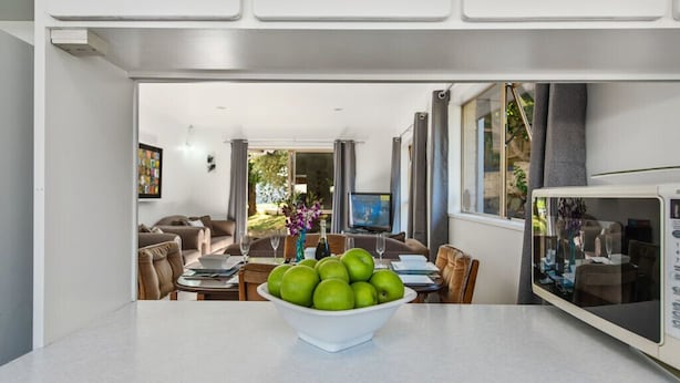 Eine moderne Küche mit einer Schüssel grüner Äpfel auf der Arbeitsfläche, ein Essbereich mit Tisch und Stühlen sowie ein Wohnzimmer mit Sofa und Fernseher.