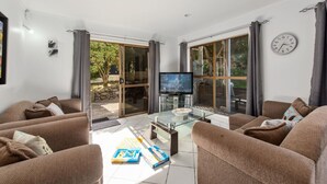 Ein Wohnzimmer mit einem Glastisch, braunen Sofas, einem Fernseher und Blick ins Freie.