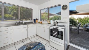 Eine moderne Küche mit weißen Schränken, einem schwarzen Backofen und einem Spülbecken. An der Wand hängt eine Uhr und es gibt ein Fenster mit Jalousien.