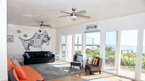 Ein geräumiges Wohnzimmer mit einer schwarzen Couch, einem grauen Sessel und einem blau gemusterten Teppich. Es gibt Deckenventilatoren, große Fenster mit Meerblick und eine Tür zu einem Außenbereich.
