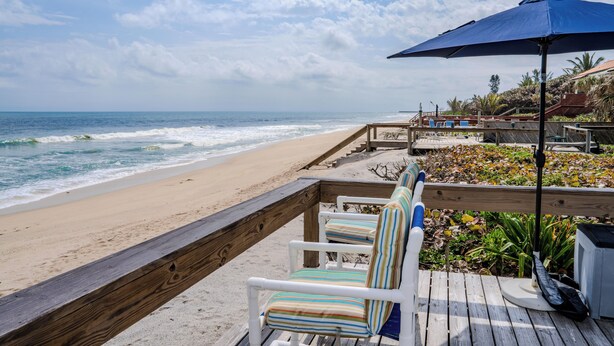 Eine Holzterrasse mit weißen Stühlen und einem blauen Sonnenschirm mit Blick auf einen Strand.