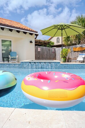 Uma piscina com um flutuador de donut inflável rosa e amarelo, um guarda-sol verde e uma cerca de madeira ao fundo.
