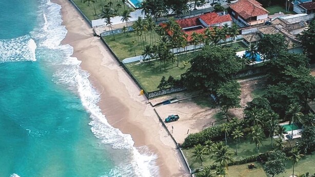 Vista aérea de una playa con aguas turquesas cristalinas, una orilla de arena y una hilera de casas con tejados rojos.