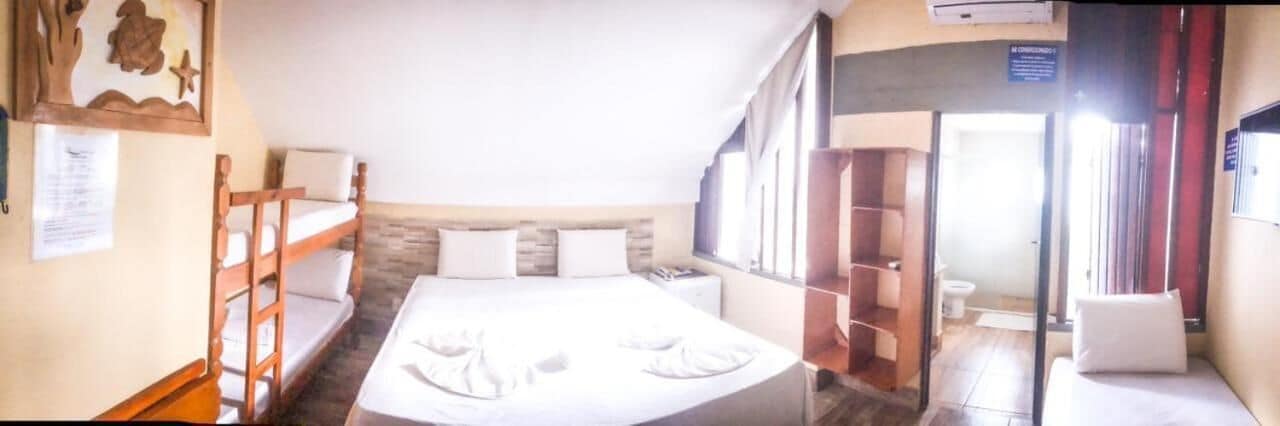 Une chambre d’hôtel avec un lit superposé, un lit simple, une tête de lit en bois, une fenêtre et une porte donnant sur une salle de bain.