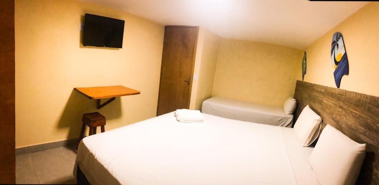 Quarto de hotel com uma cama de solteiro, uma mesinha, uma cadeira, uma televisão fixada na parede e uma porta.