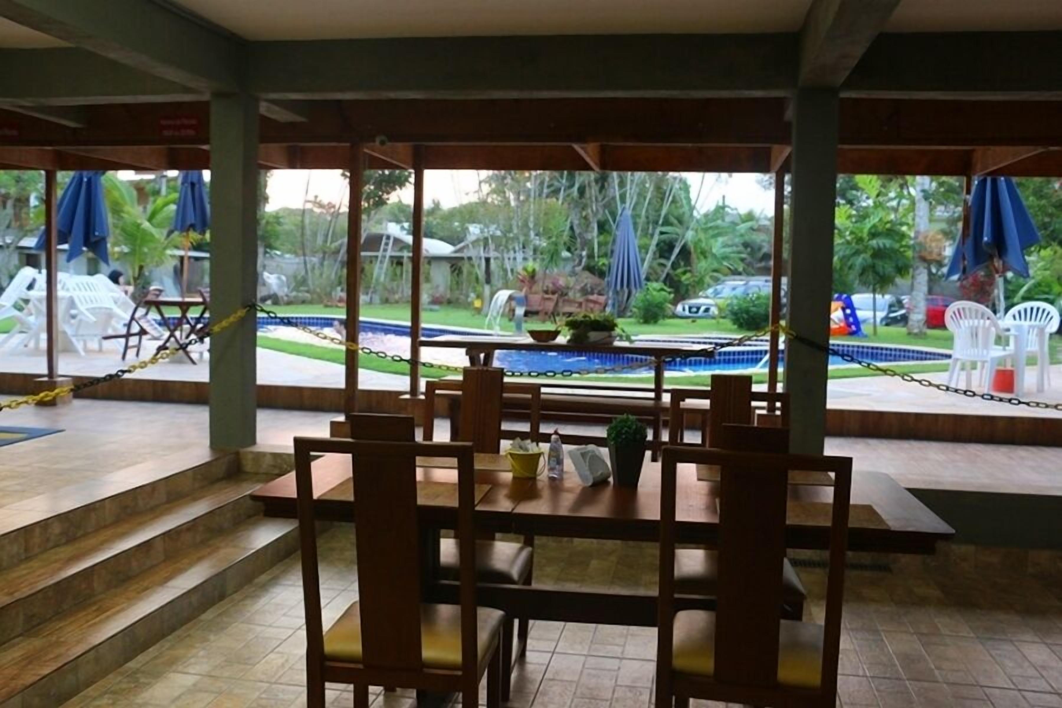 Un espacio de comedor al aire libre con mesas y sillas de madera, una piscina al fondo y vistas a un jardín.