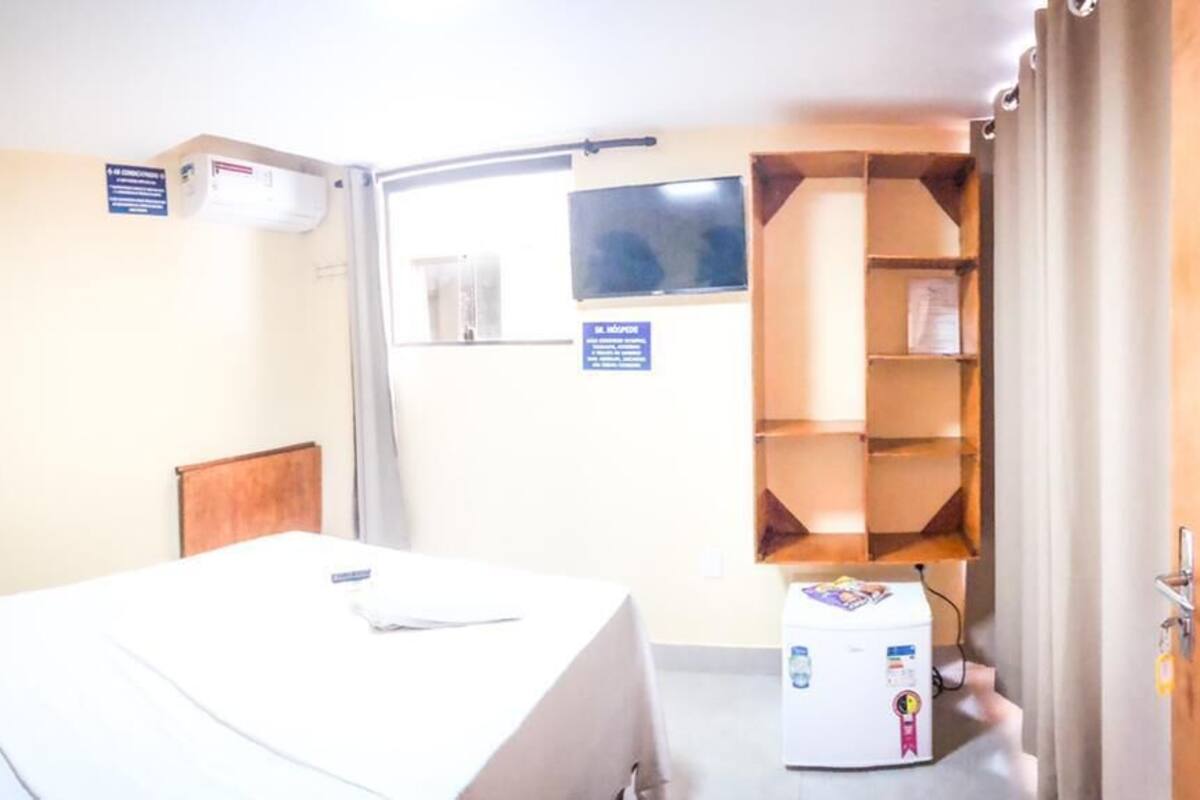 A hotel room with a bed, a small desk, a shelf with items, and a door.