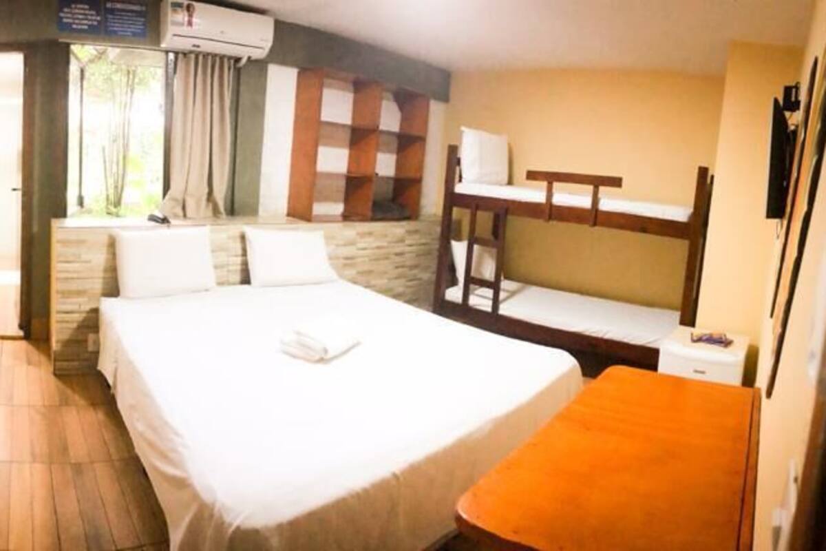 A hotel room with a bed, a window with curtains, a wooden bench, and a small table.