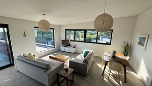 A modern living room with a sofa, coffee table, and dining area. There are large windows providing natural light and views of the outdoors. - Modern, large, open plan 1 bedroom apartment (Surfdale)