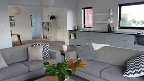 A modern living room with a grey sofa, a coffee table with a vase, and a kitchen area with white cabinets and a window. - Modern, large, open plan 1 bedroom apartment (Surfdale)