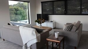 A modern living room with a grey sofa, a small coffee table, and a TV on a white cabinet. There is a large window with a view of greenery outside. - Modern, large, open plan 1 bedroom apartment (Surfdale)