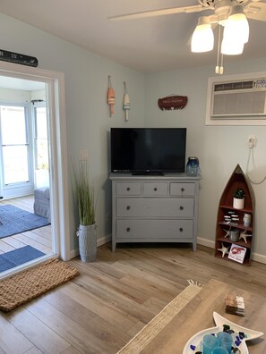 Ein Zimmer mit Flachbildfernseher auf einer grauen Kommode, Deckenventilator, Wandklimaanlage und einer kleinen Ablage mit Dekoration.