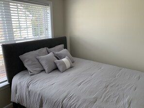 Ein Bett mit grauem Kopfteil und Kissen, ein Fenster mit Jalousien und eine schlichte Wand.