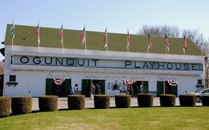 A building labeled "OGUNQUIT PLAYHOUSE" with multiple American flags on the roof, a green awning, and neatly trimmed hedges in front.