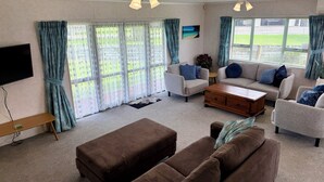 A living room with a sofa, a coffee table, and a TV. There are curtains and a window with a view of a green landscape.