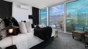 Ett modernt hotellrum med en stor säng, en svart lampa, en platt-TV och en balkong med utsikt över berg och en sjö.
