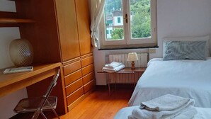 Un dormitori amb un llit, escriptori, cadira, finestra i vista cap a vegetació, i un radiador.