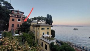 Un edifici costaner amb terrassa al terrat que fa vedar a la mar, al costat d'un edifici rosa amb balcó.