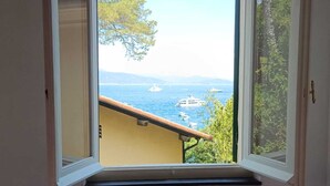 Habitació amb finestra que dona a una vista costanera, incloent vaixells i vegetació.