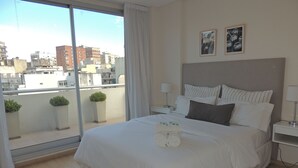 Habitación de hotel con una cama grande, vistas de la ciudad a través de una ventana, una mesita de noche con lámpara y una pequeña planta en maceta.
