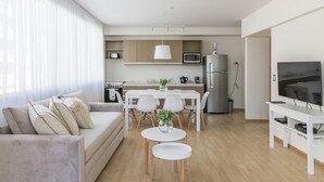 Una sala de estar moderna con un sofá, una mesa de comedor con sillas, una zona de cocina con electrodomésticos y un televisor sobre un mueble.