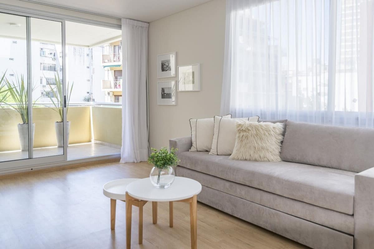 Una sala de estar moderna con un sofá gris, una mesita blanca con un florero y un ventanal con cortinas de gasa.