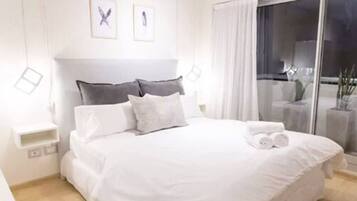 Una cama bien hecha con ropa blanca y almohadas, una mesita de noche y vistas al exterior a través de una puerta de cristal.
