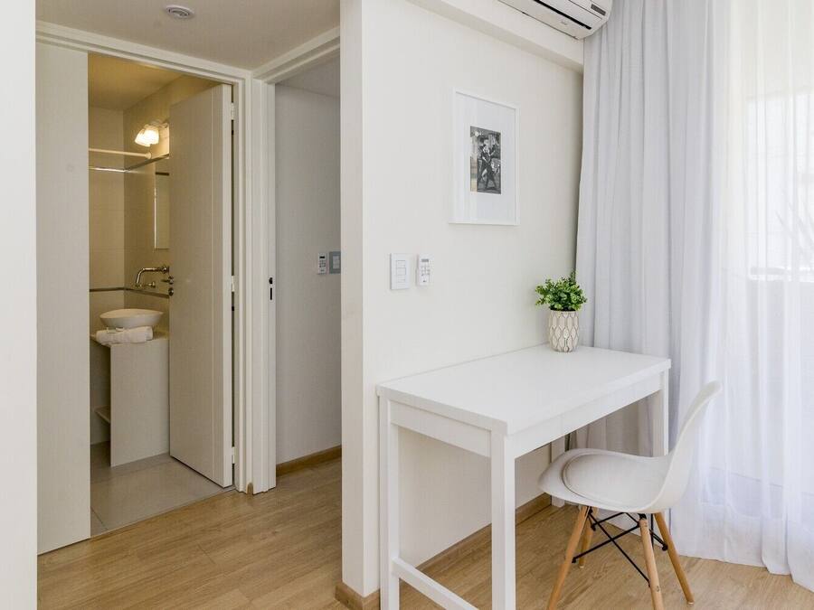 Una habitación moderna y minimalista con un escritorio blanco, una silla, una pequeña planta en maceta y un cuadro en la pared.