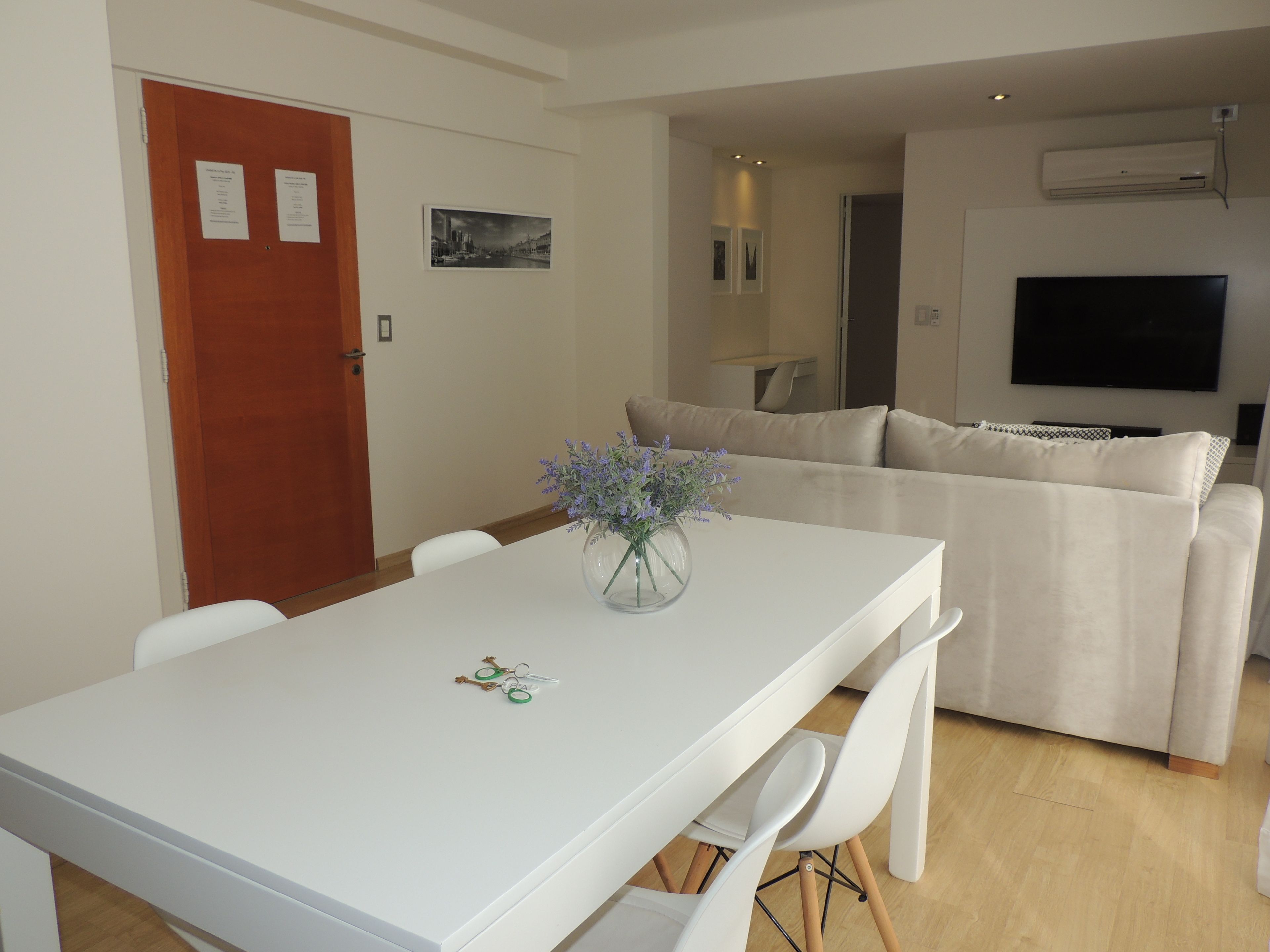 A modern living room with a white sofa, a dining table with chairs, a vase with flowers, and a flat-screen TV mounted on the wall.