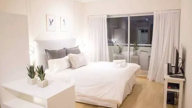 Una habitación de hotel moderna con una cama grande, una mesita de noche, un televisor y vistas al exterior a través de una puerta corrediza de vidrio.