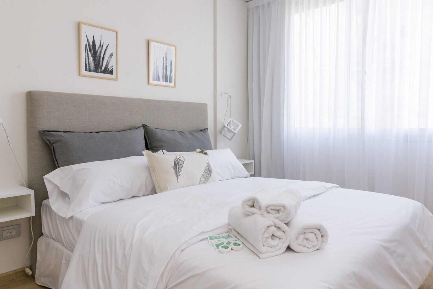 A neatly made bed with white linens and rolled towels, a grey headboard, and two framed pictures on the wall.