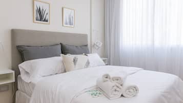 Una cama bien tendida con sábanas blancas y toallas enrolladas, un cabecero gris y dos cuadros en la pared.