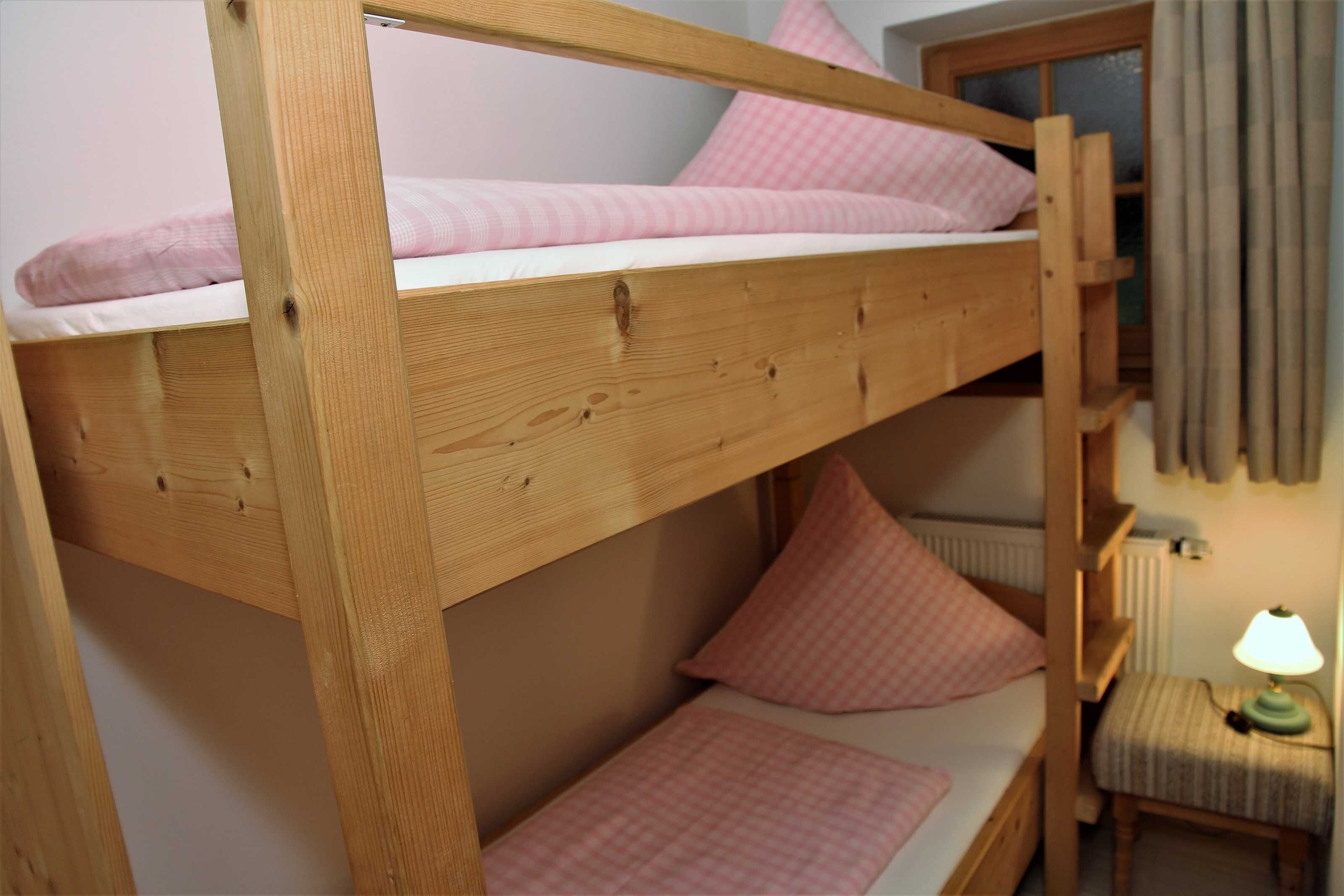 Een stapelbed met roze beddengoed, een houten hoofdbord, een klein nachtkastje met een lamp en een raam met gordijnen.