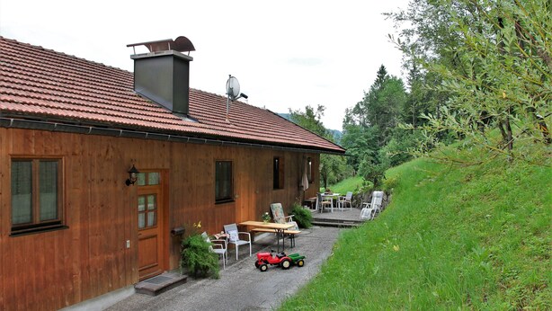 A wooden house with a red-tiled roof, a small porch with chairs, and a toy car on the ground. - Apartment 2 - Bocksleitnerhof (Wackersberg)