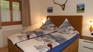 A bedroom with a bed, two bedside lamps, a window with curtains, a radiator, and wall decorations including a deer head and two framed pictures. - Apartment 2 - Bocksleitnerhof (Wackersberg)