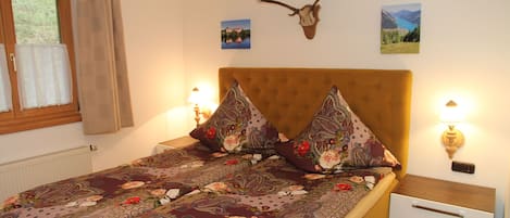 Un dormitori amb un llençol estampat amb motius florals, un panell de fusta al capçal, dues làmpades de nit, una tauleta de nit amb calaixos, una finestra amb cortines i un crani de cérvol fixat a la paret.