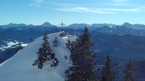 Un sommet montagneux enneigé, surmonté d’une croix, entouré d’arbres à feuilles persistantes et dominé par des montagnes lointaines, le tout sous un ciel dégagé.