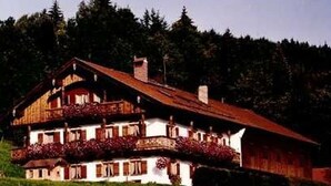 A two-story wooden house with a balcony, surrounded by greenery and trees. - Apartment 2 - Bocksleitnerhof (Wackersberg)
