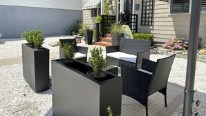 Un espace extérieur aménagé en terrasse, avec des meubles en rotin noir, des jardinières rectangulaires ornées de plantes vertes et un grand parasol noir offrant de l’ombre.