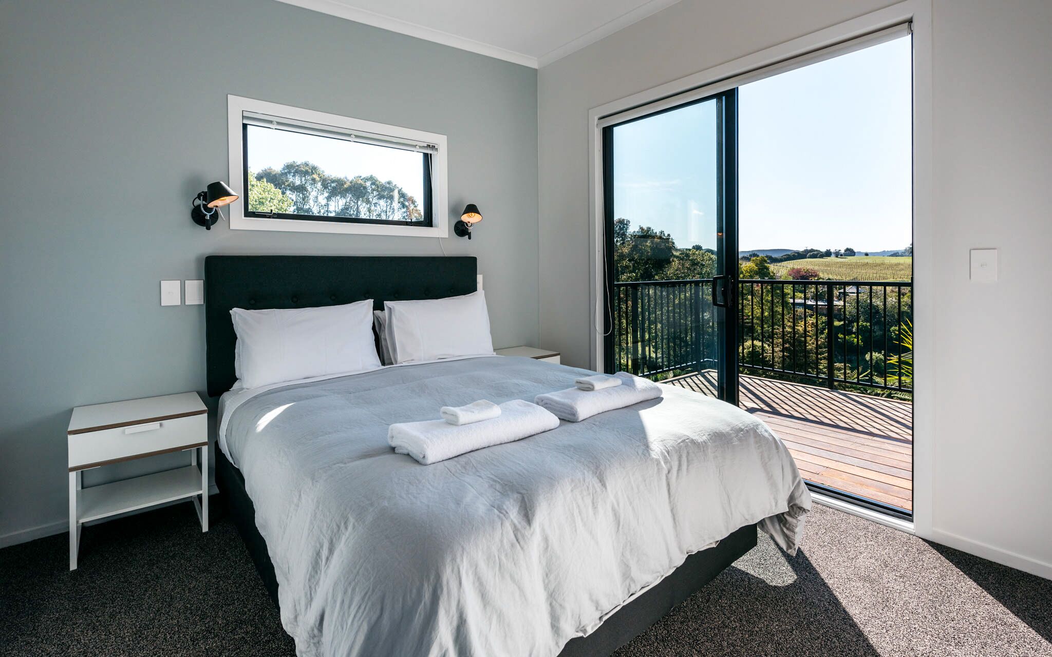 A modern bedroom with a large bed, a nightstand, a window with a view of the countryside, and a balcony with a railing.