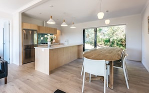 Cozinha moderna com armários de madeira, geladeira de aço inoxidável e ilha central com pia. Há uma área de jantar com mesa de madeira e cadeiras brancas.