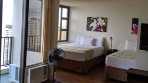 Una habitación de hotel con una cama, una mesita de noche, un ventilador, una ventana y vistas a edificios.
