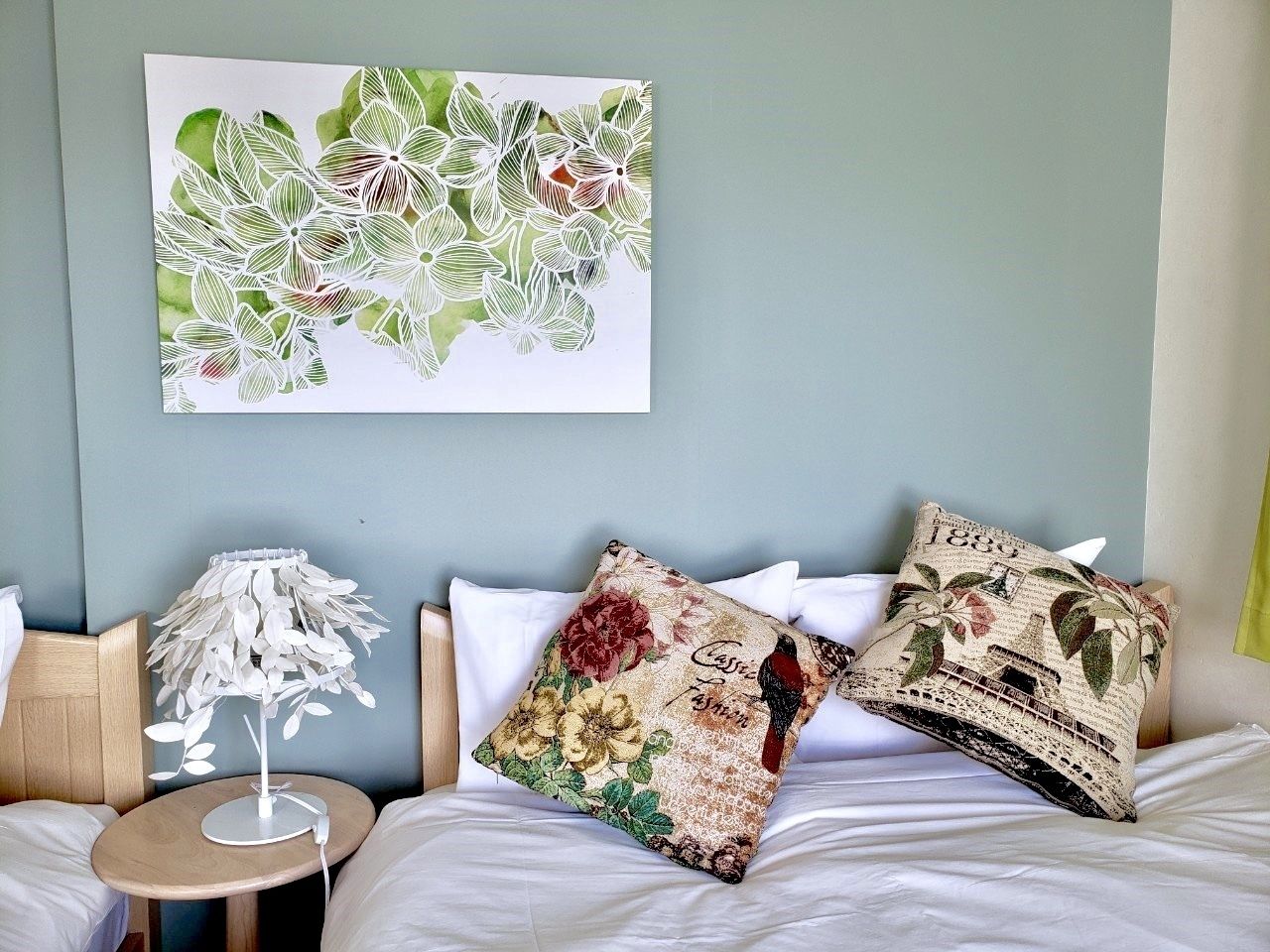 A hotel room with a bed, two pillows, a small table with a lamp, and a wall-mounted artwork.