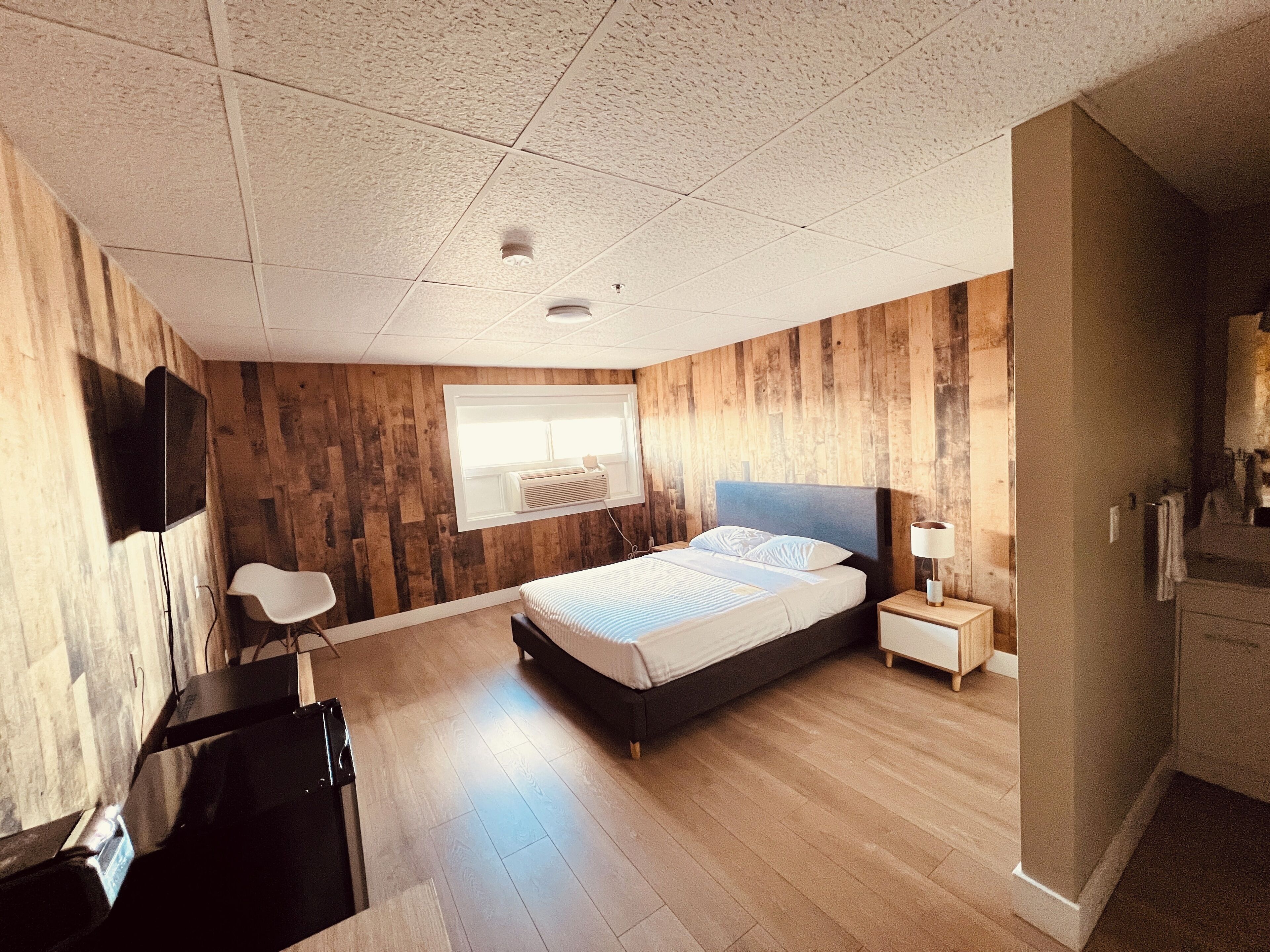 Un dormitorio moderno con panel de madera, una cama, una mesita de noche con lámpara, un televisor montado en la pared y un escritorio con silla.