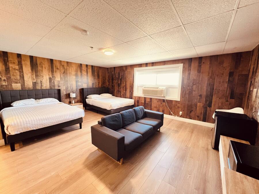 Une chambre d’hôtel avec deux lits, un canapé, des murs recouverts de panneaux de bois, un plafond doté d’un éclairage encastré et une fenêtre avec un appareil de climatisation.