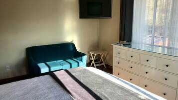 A bedroom with a bed, a teal sofa, a small table, a dresser, and a TV on the wall.
