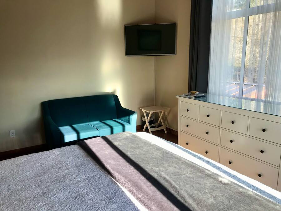A bedroom with a bed, a teal sofa, a small table, a dresser, and a TV on the wall.