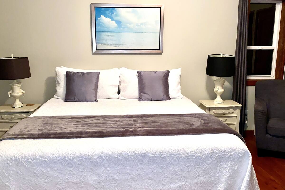 A neatly made bed with a white duvet and two pillows, flanked by bedside tables with lamps, a framed picture on the wall, and a chair in the corner.