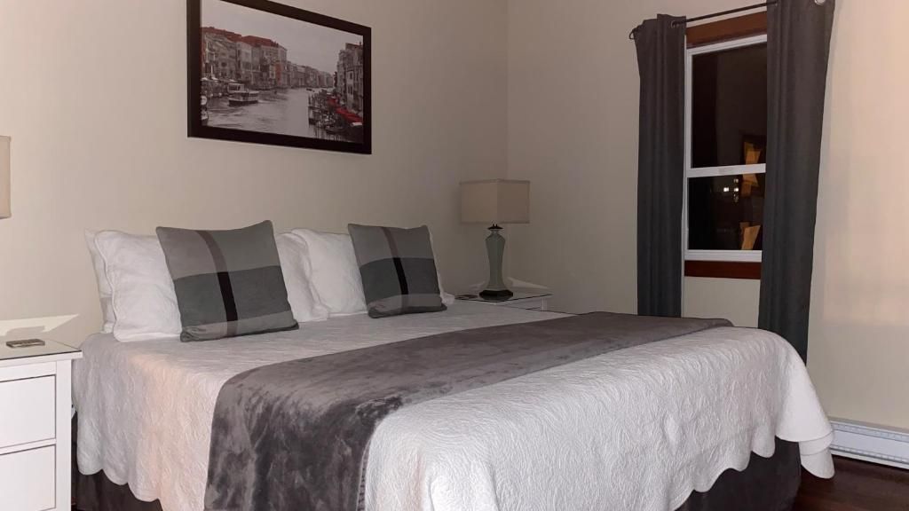 Ein Hotelzimmer mit einem Bett, einem Nachttisch, einer Lampe, einem Fenster mit Vorhängen und einem an der Wand hängenden Bild.