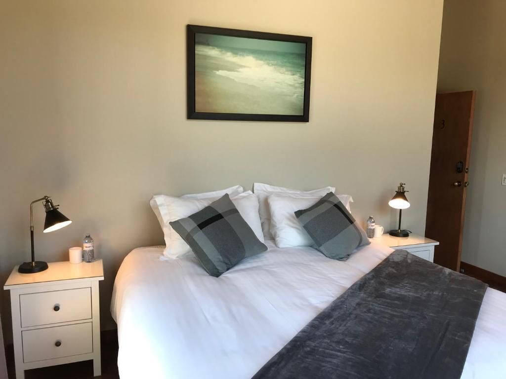 A neatly made bed with white bedding and two gray pillows, a black throw blanket, and a framed painting above it. There are two bedside tables with lamps and a small bottle on one.